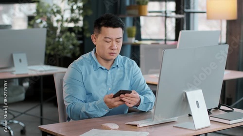 A man in a light blue shirt sits at a desk, looking intently at his smartphone. The bright office features plants and a large monitor, creating a productive atmosphere during the day.