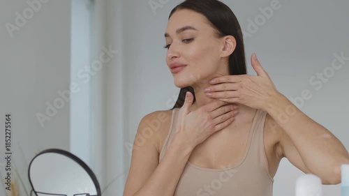 In a bright, minimalistic space, a woman gently applies skincare to her face while looking into a mirror. The natural light enhances her soft features as she engages in her beauty routine.
