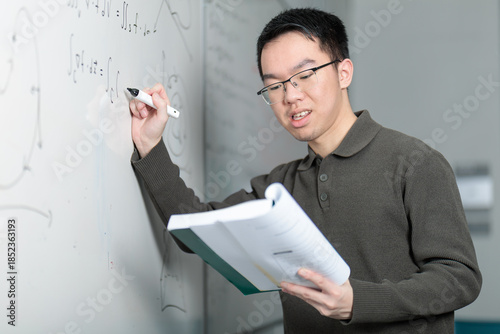 A young Chinese man writes mathematical formulas on a whiteboard with a marker while holding an open textbook. He appears focused while solving problems and teaching in a classroom setting.