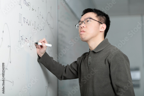 A Chinese college student wearing glasses writes mathematical equations on a whiteboard with a marker. He appears focused while working through a complex math problem in a classroom setting.