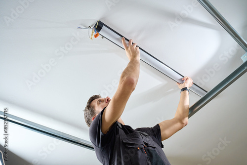 Tablou pe pânză Man in workwear installing a linear LED light fixture on the ceiling indoors