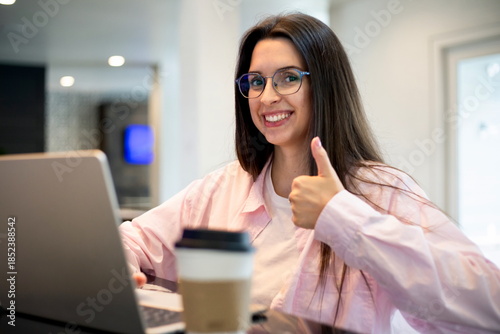 Smiling woman giving a thumbs up while working on a laptop in a bright modern interior, showing a positive and confident remote work moment.