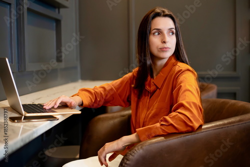 Woman seated in a modern workspace with a laptop, looking to the side, representing a professional portrait, remote work concept and contemporary digital workflow in an indoor office environment.