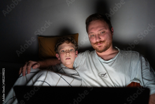 A father and son sit closely on a bed, looking at a laptop screen. The room is dark, with soft light from the laptop illuminating their faces. They are sharing a moment of laughter and joy.