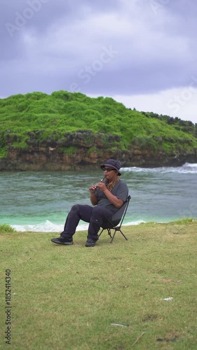 A male musician plays a tin whistle in nature with a beautiful ocean and small island in the background. A tin whistle player shows off his skills on a pristine and clean tropical beach.