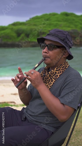 A male musician plays a tin whistle in nature with a beautiful ocean and small island in the background. A tin whistle player shows off his skills on a pristine and clean tropical beach.