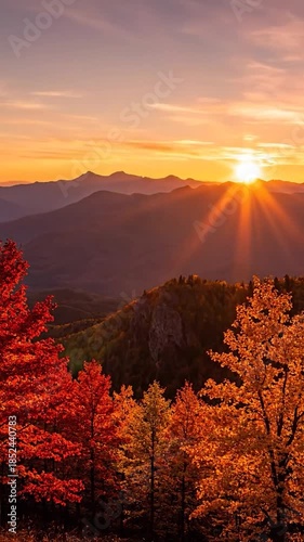 Sunset over mountain range, with vibrant autumn trees in the foreground. Sunlight bursts through the peaks