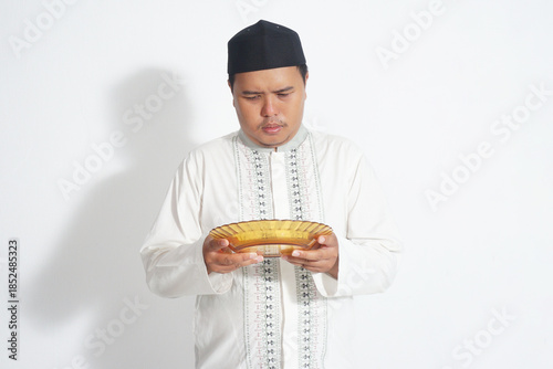 An Indonesian Muslim man wearing a black peci and a white koko shirt is holding an empty plate because he is fasting with isolated white background.
