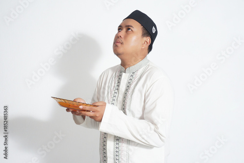 An Indonesian Muslim man wearing a black peci and a white koko shirt is holding an empty plate because he is fasting with isolated white background.