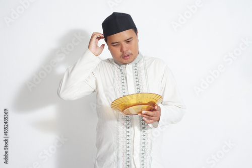 An Indonesian Muslim man wearing a black peci and a white koko shirt is holding an empty plate because he is fasting with isolated white background.