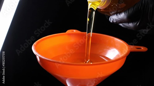 High-contrast close-up of golden amber engine oil being poured into an orange funnel, escaping the bottle's prison during routine maintenance.