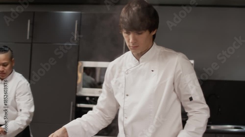 Handheld shot of male chef blanching vegetables by pouring boiling water into stainless steel bowl with tomatoes while preparing ingredients in hectic restaurant kitchen