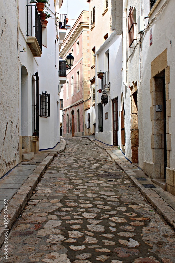Fototapeta premium Narrow medieval street with cobblestone pavement in Sitges, traditional Spanish old town architecture, white houses with balconies. Historic European alley, travel destination and charming village 