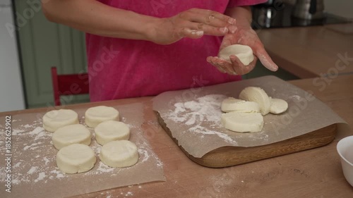 Skilled hands of a woman shaping classic cottage cheese pancakes, also known as syrniki, on a dusted countertop in a cozy kitchen before gently transferring the dough onto parchment paper for baking