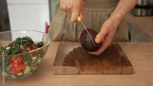 Woman's hands cutting a ripe avocado on a wooden board to prepare a healthy homemade vegetable salad in a glass bowl, showcasing a nutritious meal preparation and dieting concept