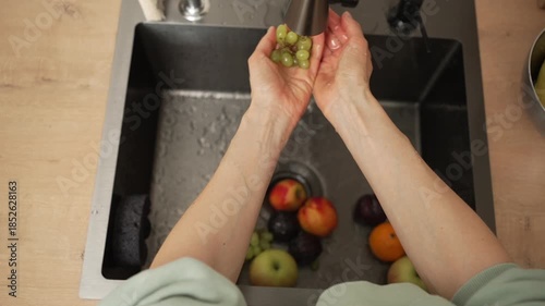Woman's hands gently rinsing a cluster of vibrant green grapes under flowing water in a contemporary kitchen sink, prepping nutritious ingredients for a delightful fruit salad at her residence