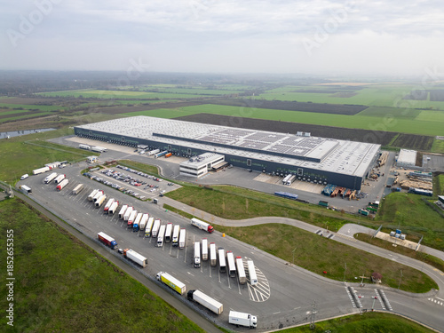 Принти на полотні Aerial view of a large logistics warehouse with loading docks and trucks, distribution center surrounded by farmland