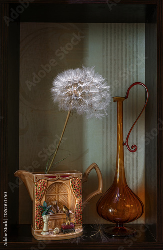 In a small niche there is a ceramic milk jug with a large fluffy dandelion and a glass pitcher.
