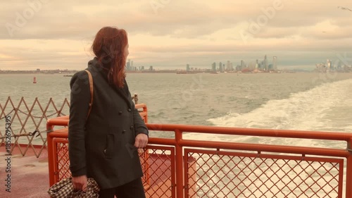 Woman with red hair looking thoughtfully at the distant Manhattan skyline New York City from the deck of a ferry during golden hour or sunset.