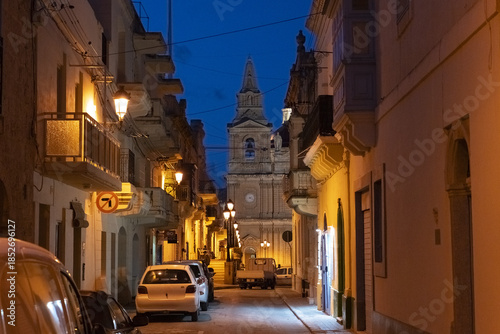 View of golden light bathing ancient stone buildings lining a narrow street leading to a towering church under a deepening blue sky, Mellieha, Malta.