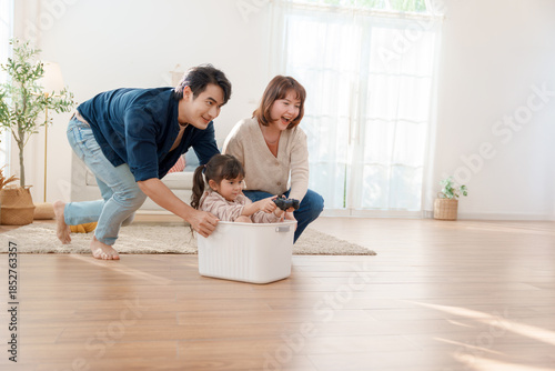 Happy Asian parents pushing their little daughter in a plastic basket while she holds a game controller at home Fun family bonding and creative childhood play in a bright modern living room
