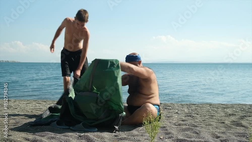 Adult overweight man and skinny teenage boy try to set up a camping tent on the seaside. Tent is complicated to assemble. Difficulty of erecting temporary shelter outdoors.