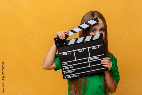 Happy little kid girl in green t-shirt holds black director film movie slate board, movie clapperboard on yellow background. Childhood, film production and human emotions concept. Mock up, copy space