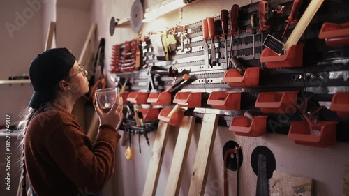 Female repairer drinking coffee and looking at tools hanging on wall in workshop interior