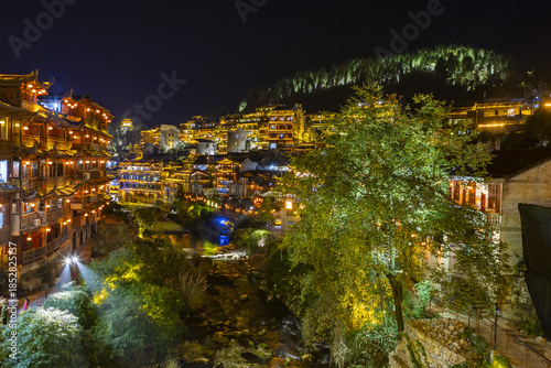 View of glowing buildings and bridges cast warm light over the dark river in a captivating panorama, Furong town, Hunan, China..