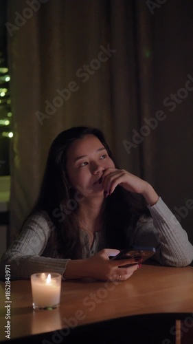A young woman sitting at a table in her cozy home during the evening, scrolling through her smartphone feed. Warm, soft lighting creates a relaxed and intimate atmosphere. Ideal for concepts of social