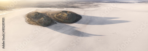 Aerial view of twin mounds cast long shadows across a vast, pale expanse, their dark forms contrasting sharply with the surrounding bleached landscape, Karynzharyk, Mangystau Region, Kazakhstan.