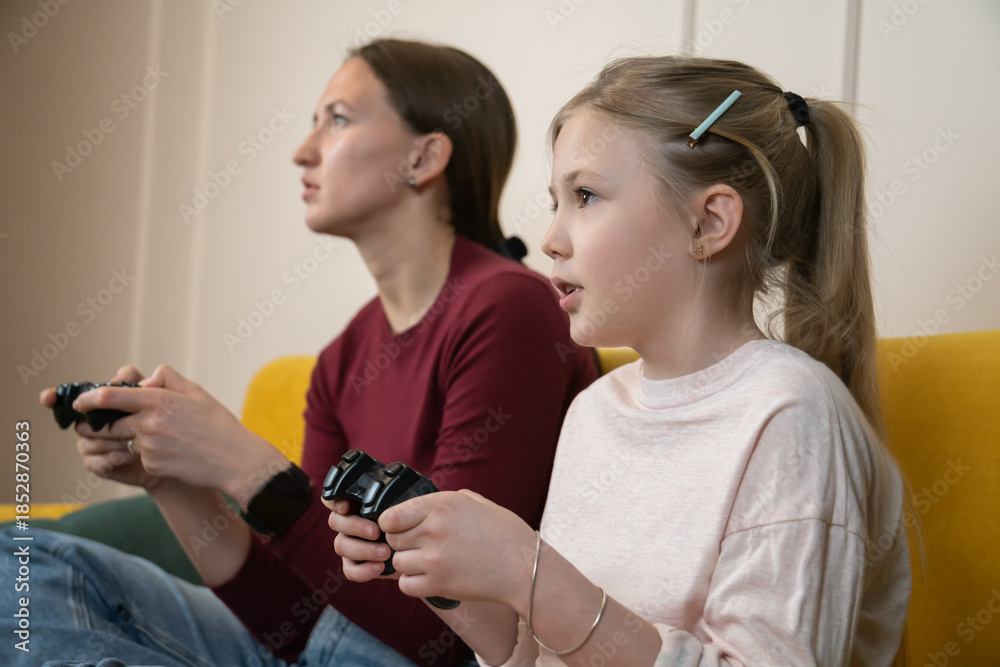 Fototapeta premium Mother and daughter sitting at home, smiling and concentrating as they play video games with controllers, sharing playful bonding, leisure and modern family entertainment