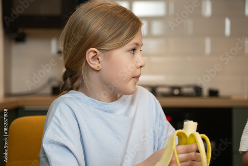 Young blonde girl eating a banana in a bright kitchen, enjoying a healthy snack that promotes balanced nutrition and happy childhood growth and natural living routines