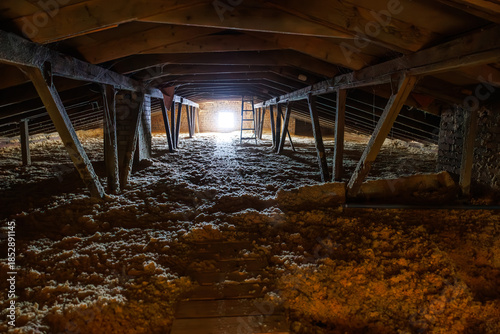 Картината върху платно Empty attic crawlspace with blown in fiberglass insulation, wooden rafters, brick pillars, and a plank walkway to a square window