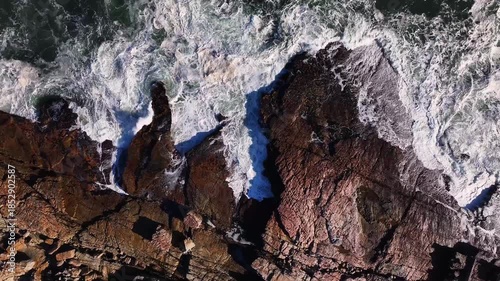 Aerial view of the rugged, rocky coastline where the churning white waves crash against the dark, textured rocks, Cape Town, Western Cape, South Africa.