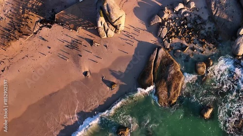 Aerial view of turquoise ocean waves crashing against the sandy beach and large boulders, creating a stunning contrast of colours, Cape Town, Western Cape, South Africa.