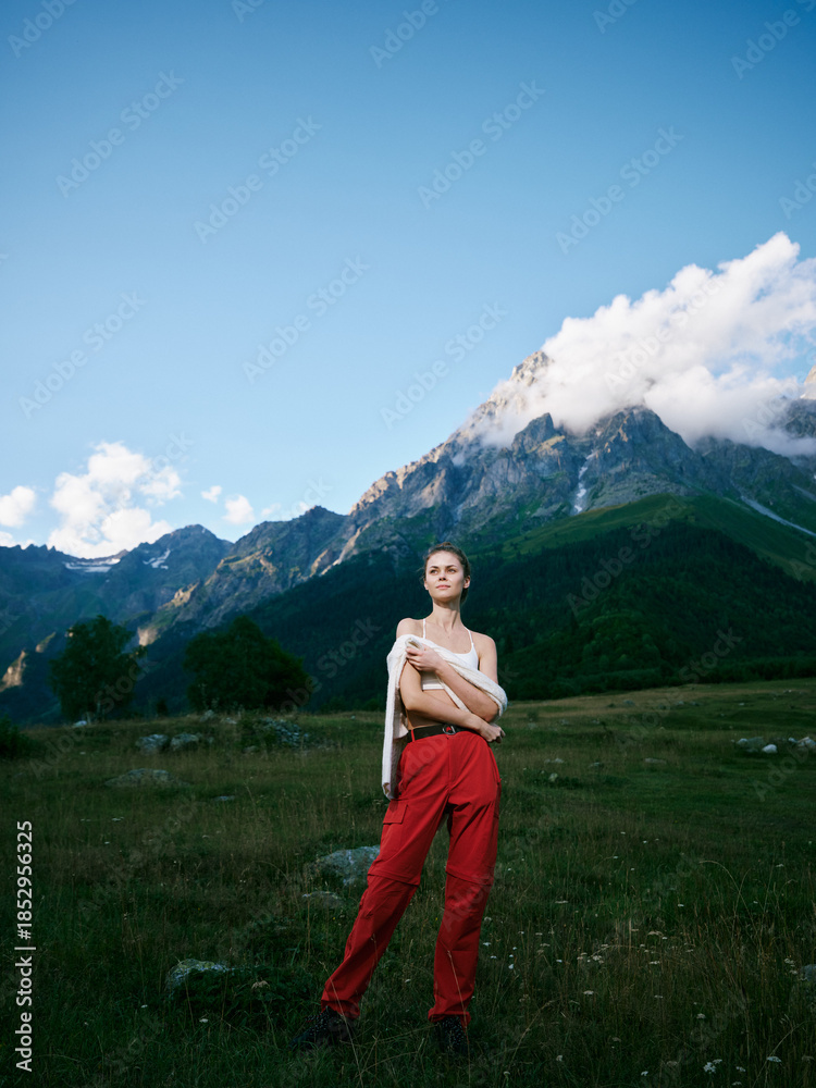 Naklejka premium Mountain landscape with a wide meadow and blue sky, a woman in red pants stands confidently, embracing outdoor adventure, nature, and serene alpine vibes in daylight