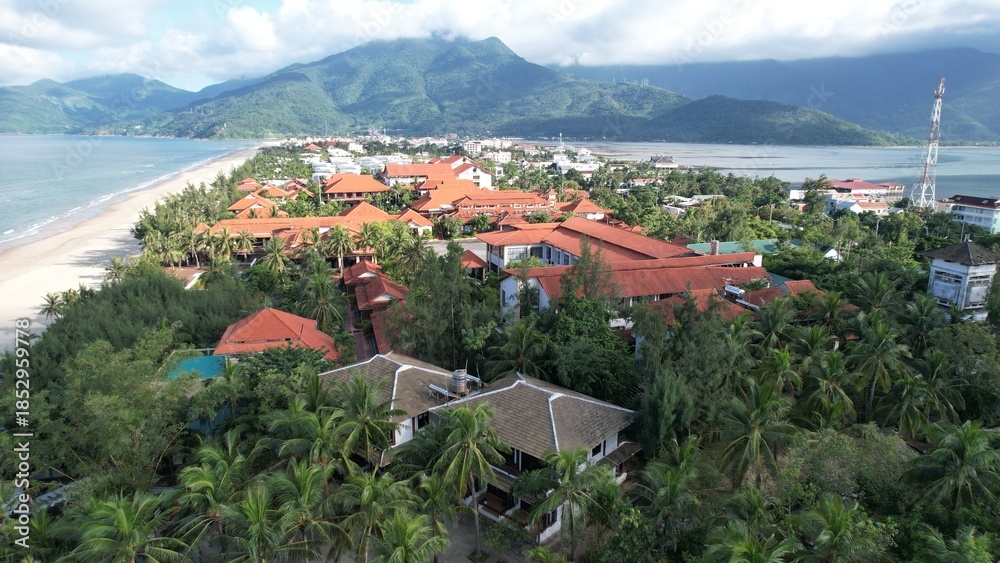 Fototapeta premium High-angle view of a coastal resort in Lang Co featuring red-tiled roofs, lush palm trees, sandy beach, and majestic mountains.