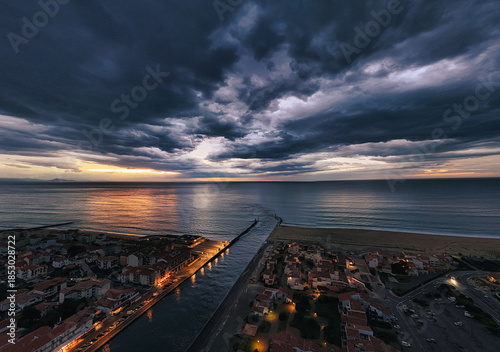 Aerial view of the canal meeting the sea under a dramatic sky, with the city lights casting a warm glow on the waters, Capbreton, France.