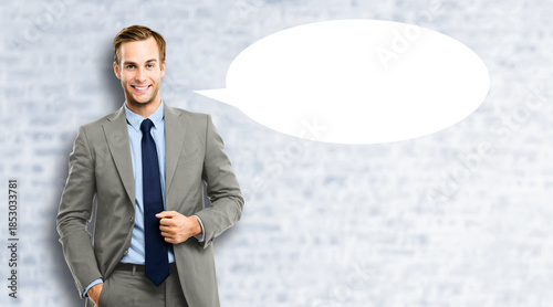 Portrait image - smile young business man wear grey gray suit jacket, blue shirt, tie. Businessman standing against bricks wall background with white empty mockup speech bubble