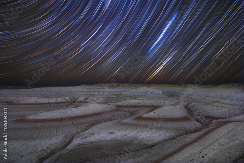 View of celestial trails dance above the arid landscape, painting the night sky with streaks of light over the red and white textured ground, Senek, Mangystau Region, Kazakhstan.