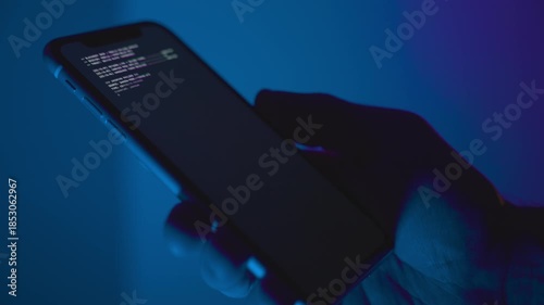 Hand holding smart phone on dark blue background while malware app is installed. Close up on device hacking code running on screen downloading and setup of illegal criminal spyware cybersecurity virus