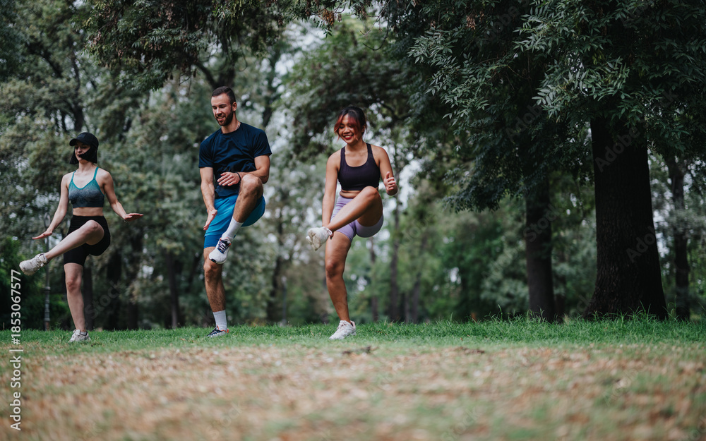 Fototapeta premium Three friends exercise in a park, lifting knees in a lively outdoor workout. They move together among trees and grass, showcasing fitness and camaraderie.