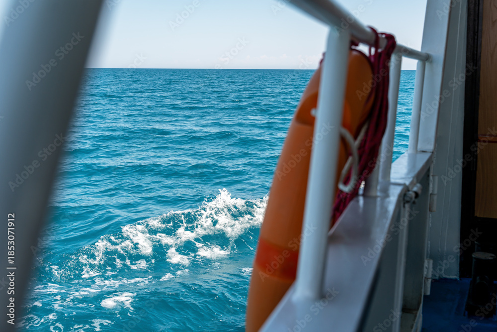Fototapeta premium A view from a boat or ferry of a large body of water with mountains in the distance and a clear blue sky. A metal railing and an orange lifebuoy are visible in the foreground.