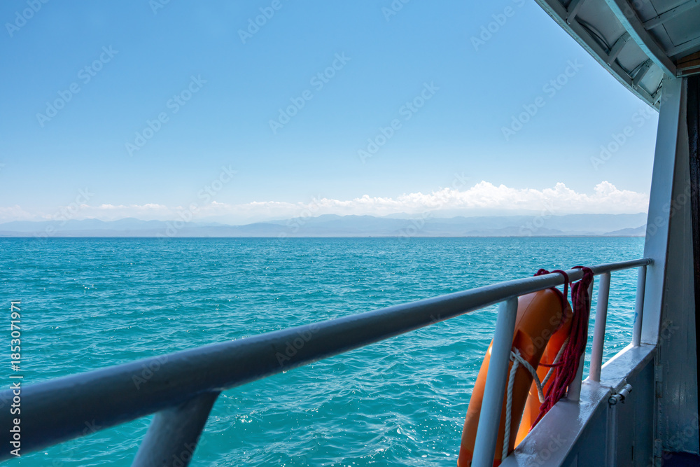 Fototapeta premium A view from a boat or ferry of a large body of water with mountains in the distance and a clear blue sky. A metal railing and an orange lifebuoy are visible in the foreground.
