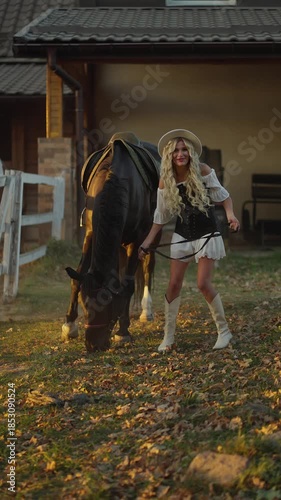 Smiling cowgirl leading a grazing horse near a countryside stable at golden hour, joyful rural lifestyle moment