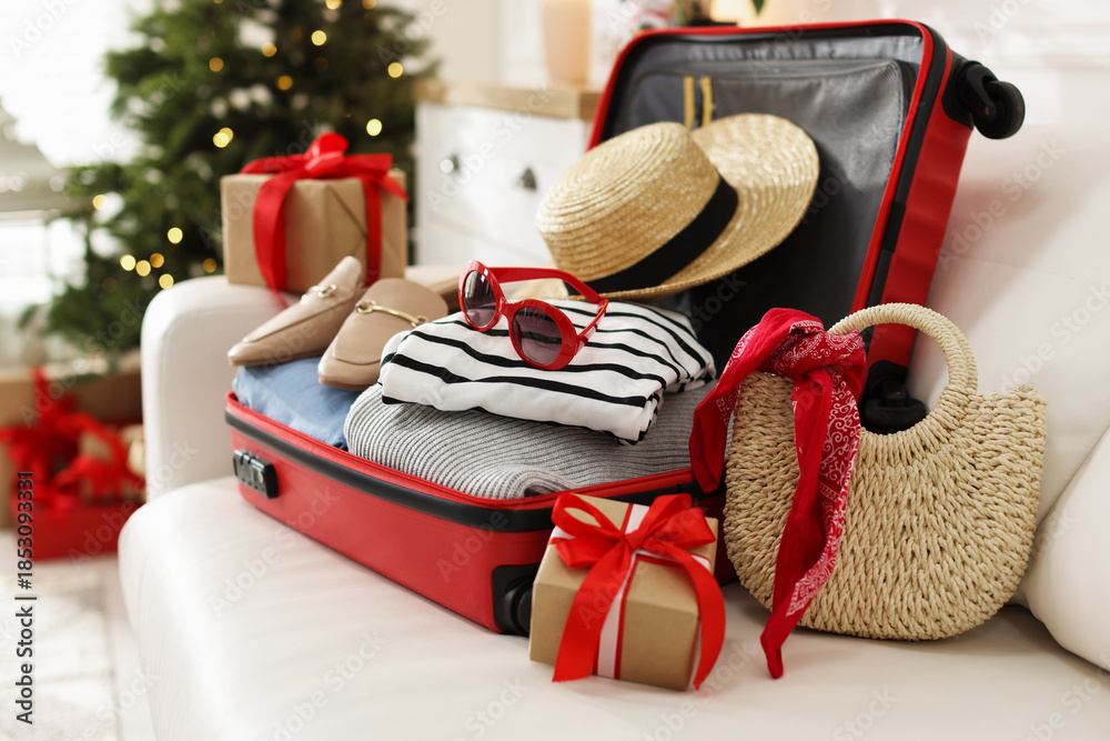 Fototapeta premium Suitcase with clothes, gifts and other belongings on sofa in festive decorated room, closeup. Christmas vacation