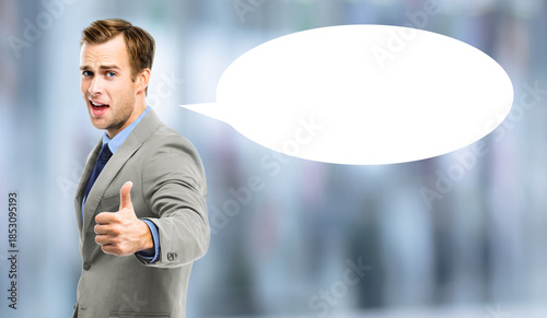 Portrait image - expressive business man in grey gray suit jacket, tie showing pointing thumb up hand sign gesture. Businessman stand  blurred office background with white empty mockup speech bubble