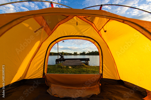A cozy yellow camping tent pitched on lush green grass by a peaceful lakeside during golden hour. nature travel, and serene outdoor living. Ideal for themes of camping, weekend getaway,