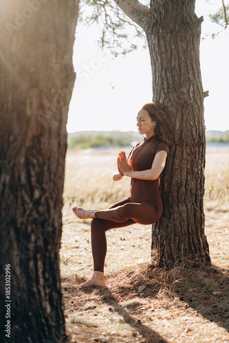 A woman practices a yoga balance pose outdoors, leaning against a tree in a calm, sunlit natural setting.